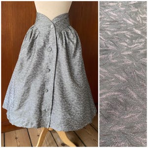 May include: A vintage grey skirt with a high, button-up waist and a subtle, textured pattern. The skirt has a flared silhouette and is displayed on a mannequin. The fabric has a slight sheen, adding to its vintage appeal.