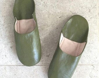 May include: Two olive green leather slippers are displayed on a light gray surface. The slippers have a rounded toe and a soft, light-colored interior. The leather appears smooth and slightly glossy.