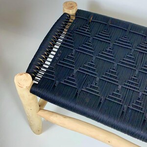 Puede incluir: Banco artesanal con asiento negro tejido y estructura de madera natural. El asiento presenta un patrón geométrico. Las patas y los soportes son de madera clara, creando un contraste con el asiento oscuro.