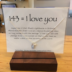 May include: A silver necklace with a charm that says "143" and a small pearl bead. The necklace is displayed on a card with the text "143 = I love you" and a description about the Minots Lighthouse in Scituate, Massachusetts. Sterling Silver - Scituate Handcrafted Jewelry by Susan Wilson.