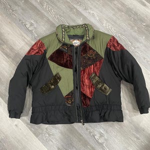 May include: A zip-up jacket featuring a color-blocked design with olive green, black, and deep red panels. The jacket includes textured velvet-like sections, patterned fabric inserts, a high collar with decorative trim, and two front pockets.