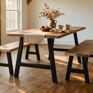 May include: A rectangular dining table with a natural wood top and black metal A-frame legs, accompanied by two matching wooden benches with black metal legs. A beige table runner, a wooden bowl with fruit, and two mugs sit on the table.