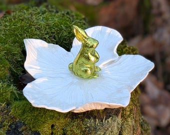 May include: A gold-colored bunny figurine sits atop a large, white, flower-shaped dish. The bunny is detailed with visible ears and a seated pose. The dish has a textured surface. The background is blurred, with green moss and brown wood visible.