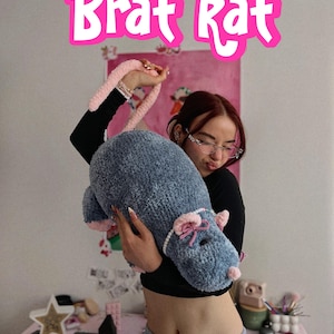 Puede incluir: Un gran cojín de felpa azul con forma de rata con detalles rosas y un asa rosa. El texto "Brat Rat" se muestra en una fuente rosa de burbujas encima del cojín.