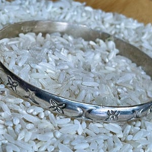 May include: A silver-toned metal bangle bracelet featuring a repeating embossed pattern of small, four-petaled flower motifs along the outer surface. The bracelet has a smooth, polished finish and a circular shape, showcasing a consistent metallic sheen.