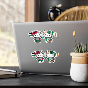 May include: Two Hello Kitty characters holding flags. The character on the left is wearing a Palestinian keffiyeh and holding a Palestinian flag. The character on the right is wearing a green shamrock and holding an Irish flag.