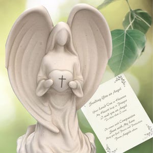 May include: White ceramic angel figurine holding a heart with a cross on it. The angel is sitting on a base and has wings. The figurine is accompanied by a card with the text "Sending You an Angel" and a poem about sending an angel to show that someone cares.