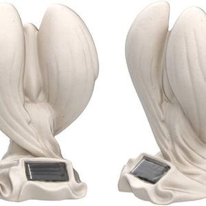 May include: White ceramic angel statue with wings outstretched, holding a small child in its arms. The statue has a solar panel on the base and is designed for outdoor use.