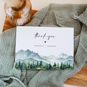 Op de afbeelding: Een witte kaart met de woorden "thank you" in elegant schrift, een klein hartje en de namen "Kendall + Jackson". De kaart toont een aquarel landschap van bergen en bomen in blauw- en groentinten.