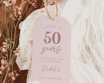 Puede incluir: Una etiqueta de regalo rosa con el texto "Cheers to 50 years" y "Thank you for celebrating Rachel's 50th Birthday". La etiqueta está suspendida por una cuerda de hilo natural. Flores rosas suaves y una tela blanca en el fondo.