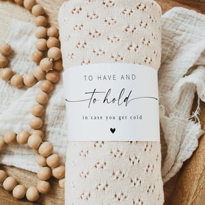 May include: A rolled, cream-coloured knitted blanket with a decorative openwork pattern. A white band wrapped around the blanket reads "TO HAVE AND to hold" and "in case you get cold" with a small heart. Wooden beads and a white cloth are in the background.