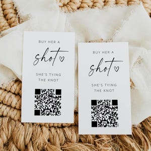 May include: Two white cards with black text that reads "BUY HER A shot SHE'S TYING THE KNOT" and a QR code. The cards are on a woven surface with a white fabric overlay.