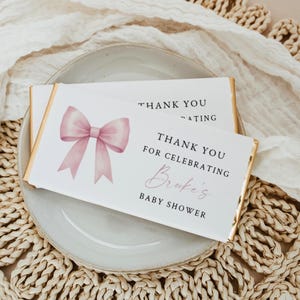 Può includere: Due incarti rettangolari per barrette di cioccolato con sfondo bianco e bordi in lamina d'oro. Ognuno presenta un fiocco rosa in stile acquerello sulla sinistra e testo nero: THANK YOU FOR CELEBRATING Brooke's BABY SHOWER.