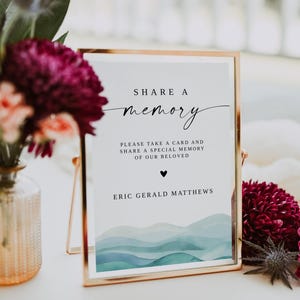 May include: A gold-framed sign with the words "SHARE A memory" in elegant script. The sign includes the text "PLEASE TAKE A CARD AND SHARE A SPECIAL MEMORY OF OUR BELOVED" and the name "ERIC GERALD MATTHEWS". The bottom of the sign features a watercolor design.