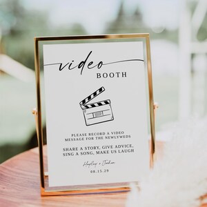 Puede incluir: Un letrero con marco dorado con las palabras "video BOOTH" en escritura elegante. Debajo, una ilustración de claqueta de cine y texto que invita a los invitados a grabar mensajes de video para los recién casados. El letrero incluye los nombres "Hayley & Jordan" y la fecha 08.15.29.