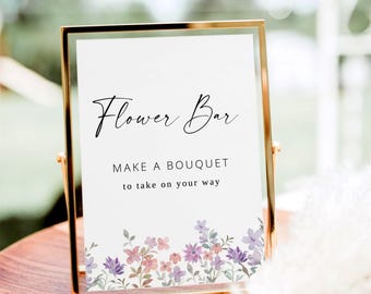 May include: A gold-framed sign with the words "Flower Bar" in elegant script. Below, it reads "MAKE A BOUQUET to take on your way." The bottom of the sign features a watercolor floral design in shades of purple and pink.