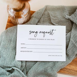 May include: A white song request card with the text "song request" in elegant script. The card includes lines for song, artist, and requestor, with the phrase "I promise to dance if you play." The card rests on a wooden board with a sage green cloth.