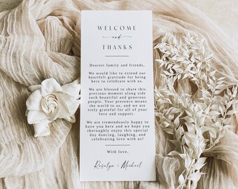 May include: A white rectangular card with the words "WELCOME and THANKS" in elegant script. The card includes a heartfelt message, surrounded by a soft, draped fabric, a white flower, and dried white floral accents. The card is a wedding or event invitation.