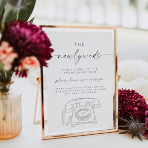 Könnte beinhalten: Ein goldgerahmtes Schild mit dem Text "THE newlyweds" und Anweisungen zum Hinterlassen einer Nachricht. Das Schild enthält eine Strichzeichnung eines Wählscheibentelefons. Das Schild ist von Blumen umgeben.