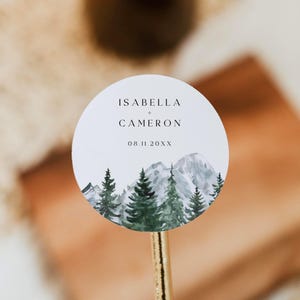 May include: A circular white sticker with the names "ISABELLA + CAMERON" and the date "08.11.20XX" printed in black. It features a watercolour illustration of a mountain range and evergreen trees in shades of green and grey.