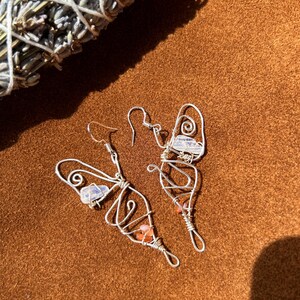 Peut inclure: Boucles d'oreilles papillon en fil d'argent, ornées de petites pierres précieuses colorées. Les boucles d'oreilles ont un aspect délicat et artisanal. Un bouquet de lavande séchée est en arrière-plan. Présentées sur une surface marron.