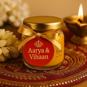 Puede incluir: Un pequeño tarro de cristal con tapa dorada y una cinta dorada atada alrededor. Una etiqueta roja con texto dorado dice "Aarya & Vihaan". El tarro está lleno de una sustancia amarilla. Una vela encendida y flores están en el fondo.