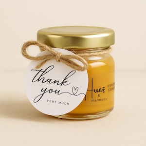 Può includere: Un piccolo barattolo di vetro con coperchio dorato, riempito di cera gialla. Un'etichetta bianca con "thank you very much" in corsivo è legata al barattolo con spago. Il barattolo ha un'etichetta con la scritta "Hues & Harmony".