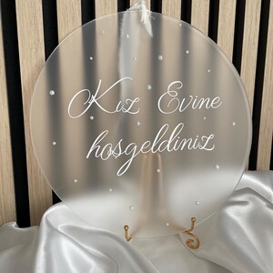 Könnte beinhalten: Ein mattes rundes Acrylschild mit dem Text "Kız Evine hoşgeldiniz" in weißen Buchstaben. Das Schild steht auf einem goldenen Ständer und hat einen weißen Satin-Stoffhintergrund.