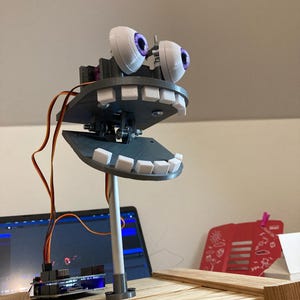 May include: A robotic face with 3D-printed white teeth and eyes with purple irises. The mouth is open, and the eyes are looking forward. The face is mounted on a white pole and connected to a circuit board with wires.