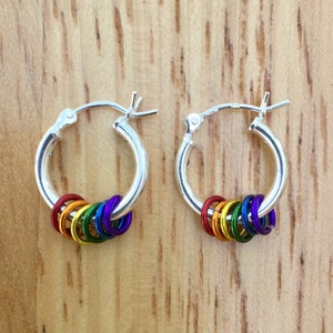 May include: A pair of silver hoop earrings, each adorned with a series of colorful rings in a rainbow spectrum. The rings are red, orange, yellow, green, blue, and purple. The earrings are displayed on a wooden surface.