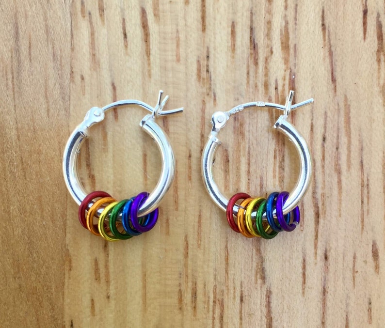 May include: A pair of silver hoop earrings, each adorned with a series of colorful rings in a rainbow spectrum. The rings are red, orange, yellow, green, blue, and purple. The earrings are displayed on a wooden surface.