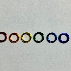 May include: Six colorful jump rings in a row, each with a small opening. The colors are red, orange, yellow, green, blue, and indigo. The rings are made of metal and have a shiny finish.