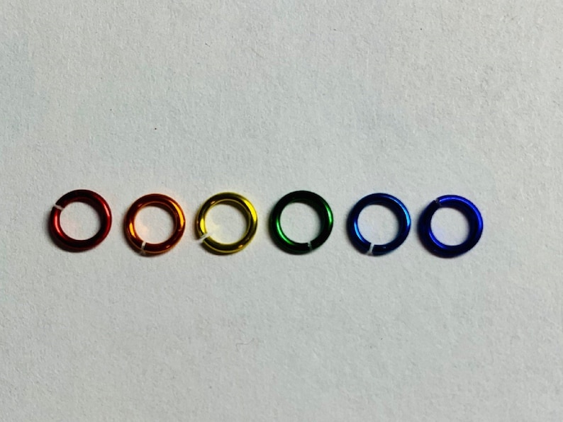 May include: Six colorful jump rings in a row, each with a small opening. The colors are red, orange, yellow, green, blue, and indigo. The rings are made of metal and have a shiny finish.