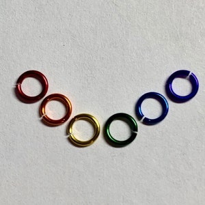 May include: A collection of six colorful metal jump rings arranged in a rainbow arc. The rings are red, orange, yellow, green, blue, and purple. Each ring is approximately 0.5 inches in diameter and has a small opening.