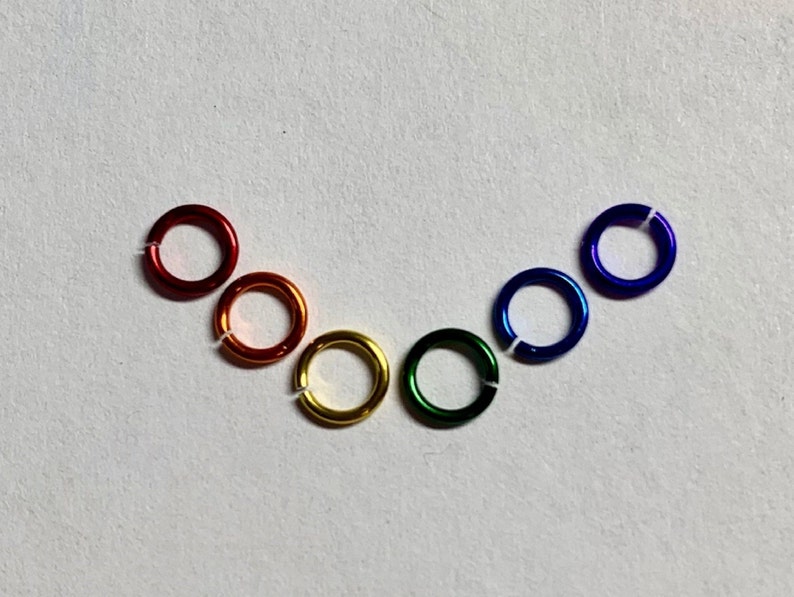 May include: A collection of six colorful metal jump rings arranged in a rainbow arc. The rings are red, orange, yellow, green, blue, and purple. Each ring is approximately 0.5 inches in diameter and has a small opening.