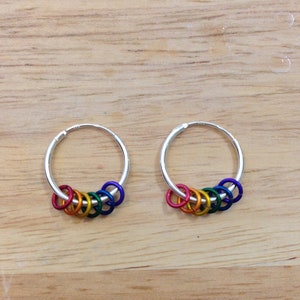 May include: A pair of silver hoop earrings, each adorned with a series of small, colorful rings in a rainbow spectrum. The earrings rest on a light brown wooden surface, showcasing the vibrant colors.