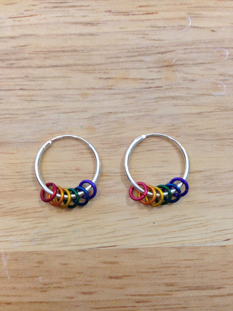 May include: A pair of silver hoop earrings, each adorned with a series of small, colorful rings in a rainbow spectrum. The earrings rest on a light brown wooden surface, showcasing the vibrant colors.