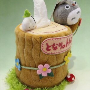 May include: A plush tissue box cover shaped like a brown tree stump with a gray Totoro character, a small white spirit, and felt flowers. The stump features a pink oval patch with Japanese text and a green grassy base with a red mushroom.