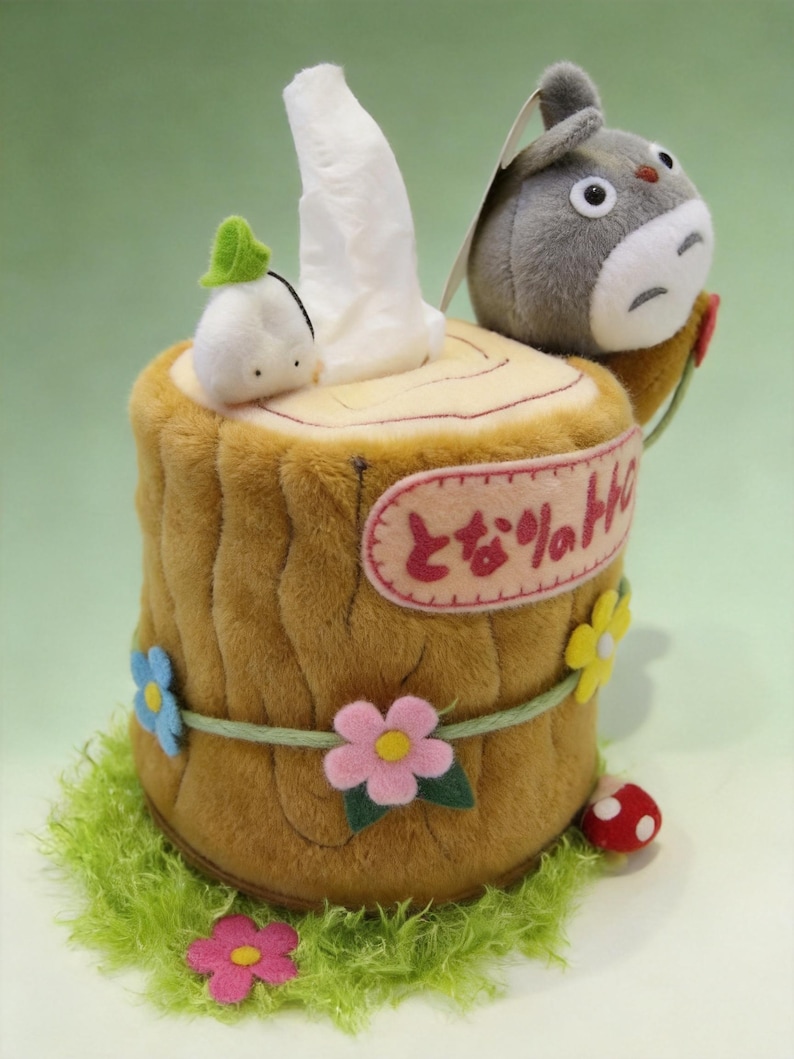 May include: A plush tissue box cover shaped like a brown tree stump with a gray Totoro character, a small white spirit, and felt flowers. The stump features a pink oval patch with Japanese text and a green grassy base with a red mushroom.