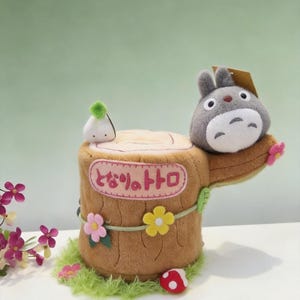 May include: A plush tree stump featuring a grey Totoro character, a small white spirit, and a red mushroom. The stump has pink, yellow, and green felt flowers, green grass trim, and a pink rectangular patch with the Japanese text となりのトトロ.