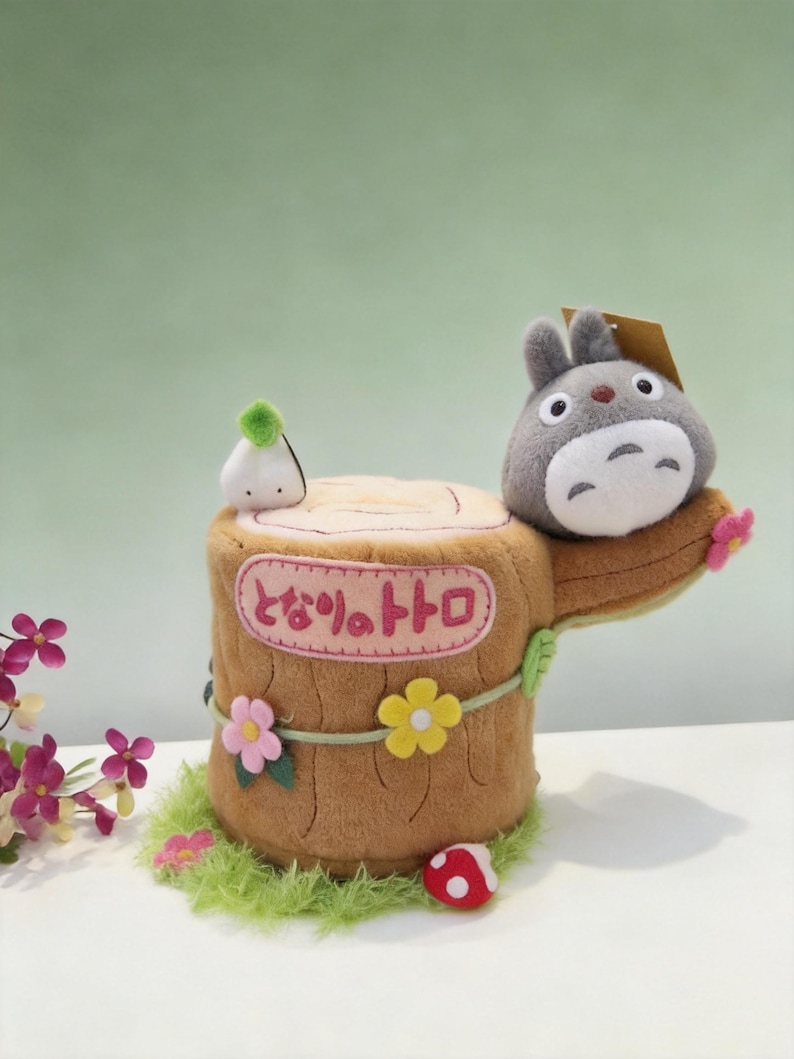 May include: A plush tree stump featuring a grey Totoro character, a small white spirit, and a red mushroom. The stump has pink, yellow, and green felt flowers, green grass trim, and a pink rectangular patch with the Japanese text となりのトトロ.