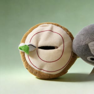 May include: A round plush toy shaped like a tree stump with a brown textured exterior and a cream-colored top featuring a red spiral pattern, a horizontal slit opening, and a small green leaf accent attached to the side next to a gray plush.