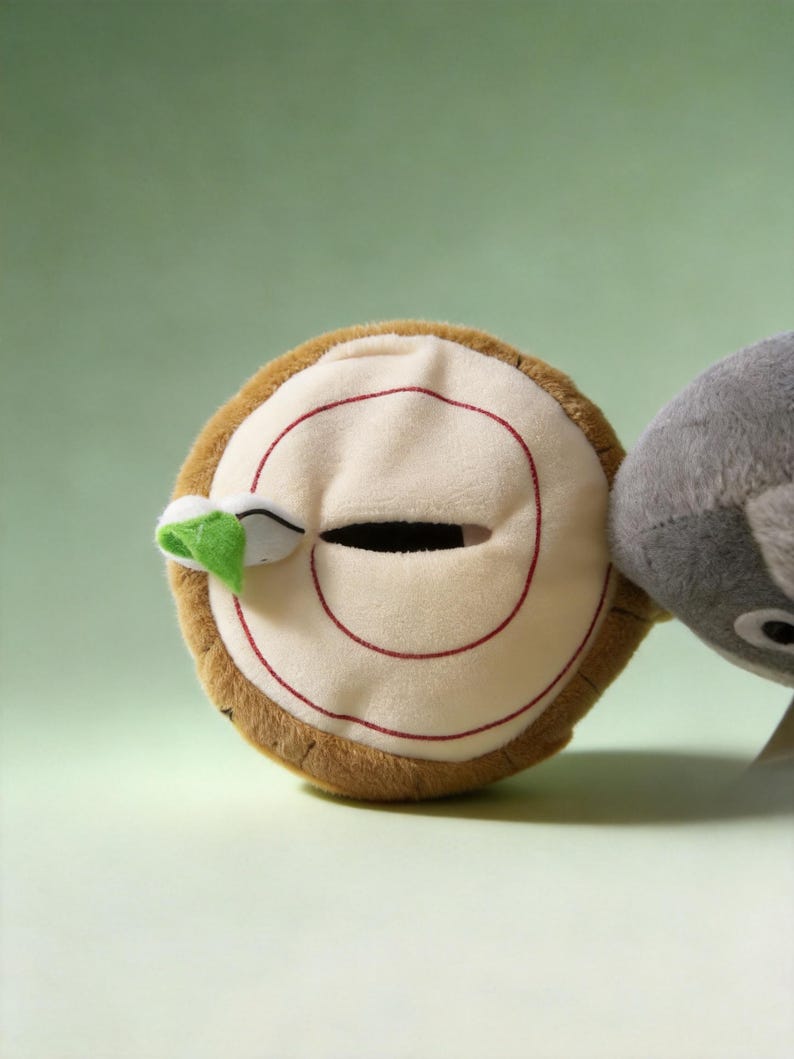 May include: A round plush toy shaped like a tree stump with a brown textured exterior and a cream-colored top featuring a red spiral pattern, a horizontal slit opening, and a small green leaf accent attached to the side next to a gray plush.