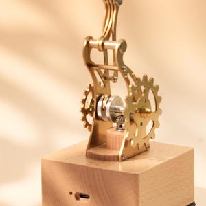 Puede incluir: Un modelo mec&aacute;nico de color lat&oacute;n con engranajes y una base de madera. El intrincado dise&ntilde;o presenta m&uacute;ltiples partes m&oacute;viles, incluyendo una serie de engranajes y un brazo giratorio. La base tiene un puerto USB-C.