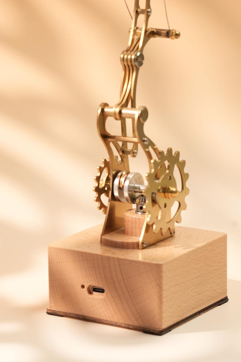 Puede incluir: Un modelo mec&aacute;nico de color lat&oacute;n con engranajes y una base de madera. El intrincado dise&ntilde;o presenta m&uacute;ltiples partes m&oacute;viles, incluyendo una serie de engranajes y un brazo giratorio. La base tiene un puerto USB-C.