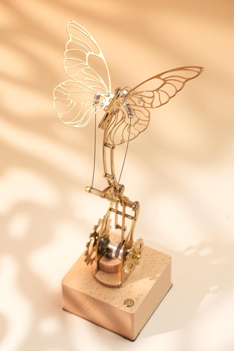 Puede incluir: Una escultura de mariposa de metal dorado con alas detalladas, engranajes y componentes mec&aacute;nicos. La mariposa est&aacute; montada sobre una base de madera, mostrando una combinaci&oacute;n de arte e ingenier&iacute;a. Las alas de la escultura miden unos 15 cm de ancho.
