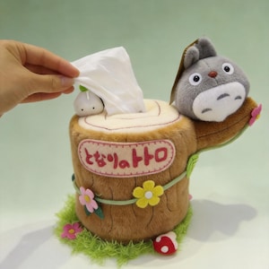 May include: A plush tissue box cover shaped like a brown tree stump with a grey character figure on top. The stump features pink, yellow, and white felt flowers, a small white mushroom, and Japanese text in a pink oval. A white tissue is being pulled out.