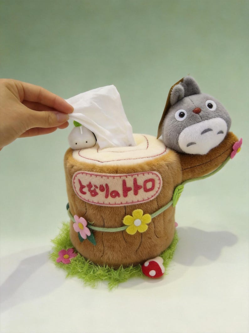 May include: A plush tissue box cover shaped like a brown tree stump with a grey character figure on top. The stump features pink, yellow, and white felt flowers, a small white mushroom, and Japanese text in a pink oval. A white tissue is being pulled out.