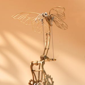 Puede incluir: Escultura mec&aacute;nica de mariposa en color lat&oacute;n, con intrincados detalles de engranajes y alas. Las alas son delicadas y transparentes, y la escultura est&aacute; montada sobre una base de madera. Los engranajes y palancas sugieren movimiento.