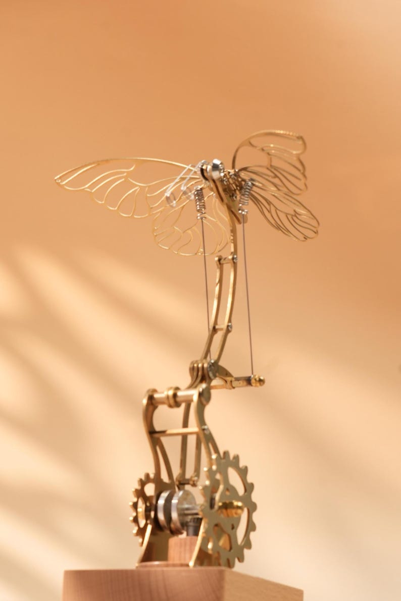 Puede incluir: Escultura mec&aacute;nica de mariposa en color lat&oacute;n, con intrincados detalles de engranajes y alas. Las alas son delicadas y transparentes, y la escultura est&aacute; montada sobre una base de madera. Los engranajes y palancas sugieren movimiento.